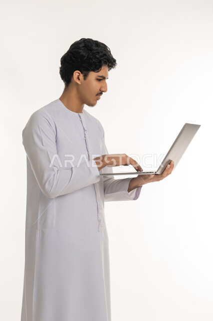 تصفح مواقع التواصل الإجتماعي، ايماءات التركيز والاندماج، مشاهدة الأفلام والمسلسلات، انجاز الاعمال المطلوبة، دمج التكنولوجيا بالحياة العملية، بورتريه لشاب عربي خليجي إماراتي يرتدي الكندورة يقف ويمسك بيده الحاسب المحمول، خلفية بيضاء