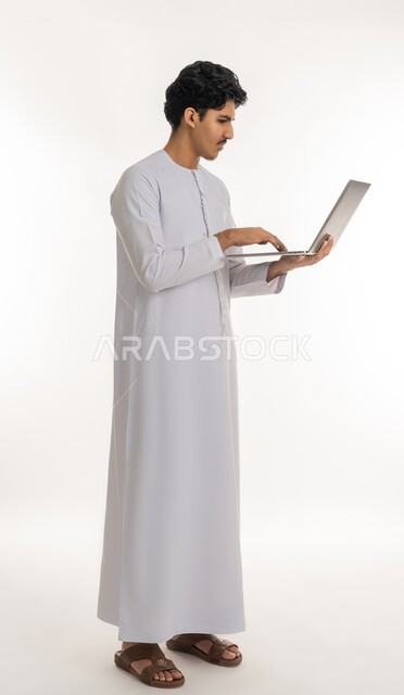دمج التكنولوجيا بالحياة العملية، بورتريه لشاب عربي خليجي إماراتي يرتدي الكندورة يقف ويمسك بيده الحاسب المحمول، تصفح مواقع التواصل الإجتماعي، ايماءات التركيز والاندماج، مشاهدة الأفلام والمسلسلات، انجاز الاعمال المطلوبة، الطول الكامل للجسم، خلفية بيضاء