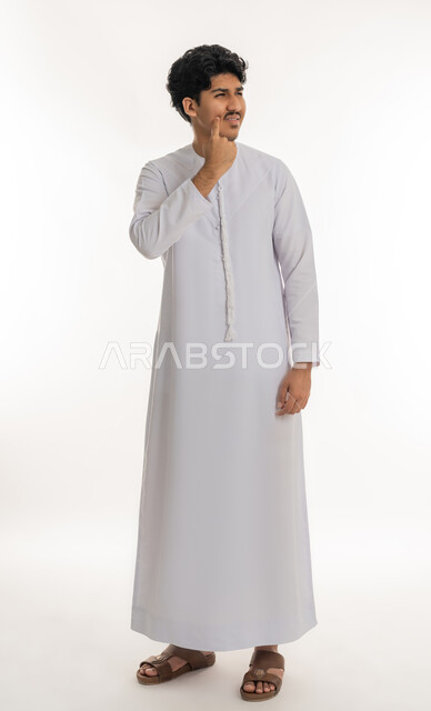Trying to find a solution to a problem, contemplation, concentration and planning for the future, portrait of a young Arab Gulf Emirati man wearing a kandura, standing and putting his hand on his cheek with expressions of thinking and contemplation, mind wandering somewhere, looking away to remember something, gestures of confusion and anxiety, full body length, white background