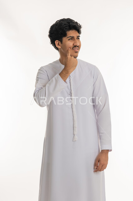 شرود الذهن إلى مكان ما, النظر بعيداً لتذكر شيء، إيماءات الحيرة والقلق, محاولة إيجاد حل مشكلة، التمعن والتركيز والتخطيط للمستقبل، بورتريه لشاب عربي خليجي إماراتي يرتدي الكندورة يقف ويضع يده على خده بتعابير التفكير والتأمل، خلفية بيضاء
