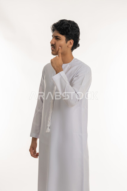 التمعن والتركيز والتخطيط للمستقبل، بورتريه لشاب عربي خليجي إماراتي يرتدي الكندورة يقف ويضع يده على خده بتعابير التفكير والتأمل، شرود الذهن إلى مكان ما, النظر بعيداً لتذكر شيء، إيماءات الحيرة والقلق, محاولة إيجاد حل مشكلة، خلفية بيضاء