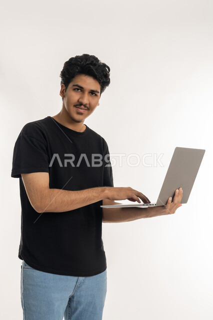 مشاهدة الأفلام والمسلسلات، مفهوم الاجتهاد وانجاز الاعمال المطلوبة، دمج التكنولوجيا بالحياة العملية، بورتريه لشاب عربي خليجي إماراتي يرتدي الزي الكاجوال ينظر الى الكاميرا ويمسك بيده الحاسب المحمول، تصفح مواقع التواصل الإجتماعي، خلفية بيضاء