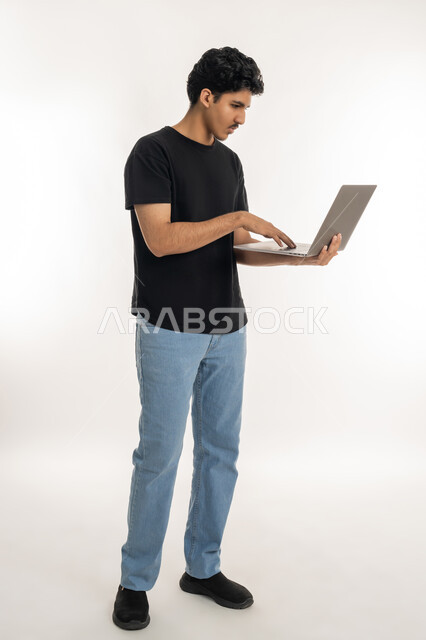 انجاز الاعمال المطلوبة، دمج التكنولوجيا بالحياة العملية، بورتريه لشاب عربي خليجي إماراتي يرتدي الزي الكاجوال يقف ويمسك بيده الحاسب المحمول، تصفح مواقع التواصل الإجتماعي، ايماءات التركيز والاندماج، مشاهدة الأفلام والمسلسلات، الطول الكامل للجسم، خلفية بيضاء