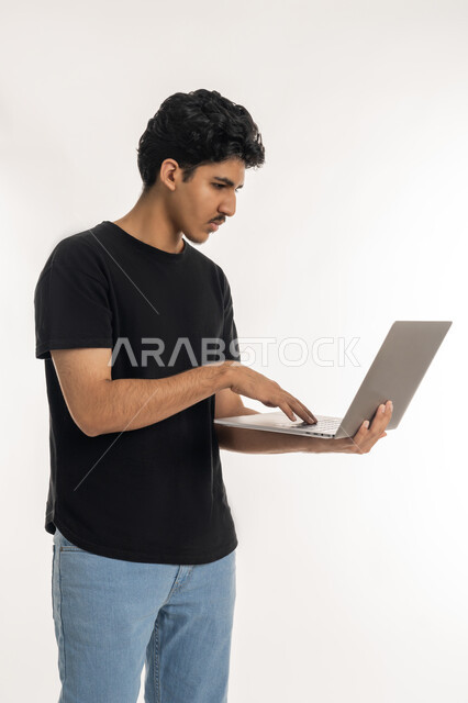 ايماءات التركيز والاندماج، مشاهدة الأفلام والمسلسلات، مفهوم الاجتهاد وانجاز الاعمال المطلوبة، دمج التكنولوجيا بالحياة العملية، بورتريه لشاب عربي خليجي إماراتي يرتدي الزي الكاجوال يقف ويمسك بيده الحاسب المحمول، تصفح مواقع التواصل الإجتماعي، خلفية بيضاء