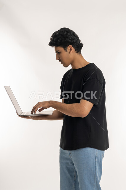 ايماءات التركيز والاندماج، مشاهدة الأفلام والمسلسلات، مفهوم الاجتهاد وانجاز الاعمال المطلوبة، دمج التكنولوجيا بالحياة العملية، بورتريه لشاب عربي خليجي إماراتي يرتدي الزي الكاجوال يقف ويمسك بيده الحاسب المحمول، تصفح مواقع التواصل الإجتماعي، خلفية بيضاء