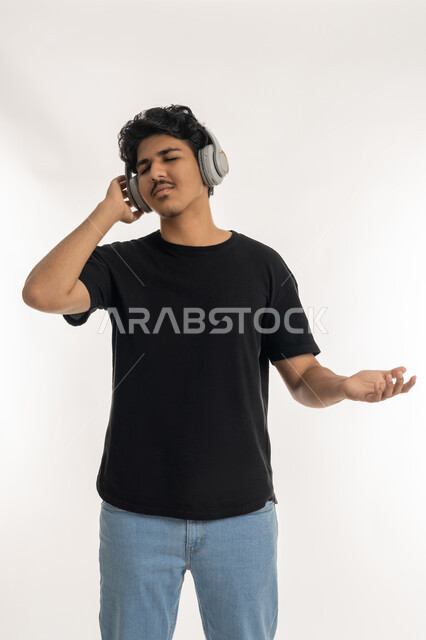 مفهوم الإصغاء باسخدام الهيدفون، بورتريه لشاب عربي خليجي إماراتي يرتدي الزي الكاجوال يضع سماعات الرأس بإيماءات الفرح والسرور، التفاعل والاندماج مع الأنغام الموسيقية, الاستماع للأغاني والمواهب الغنائية، خلفية بيضاء