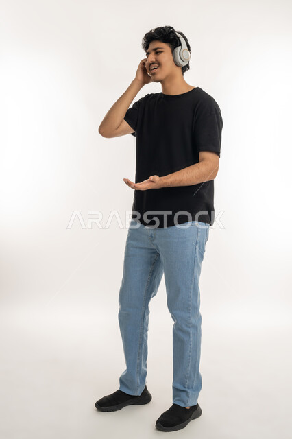 التفاعل والاندماج مع الأنغام الموسيقية, الاستماع للأغاني والمواهب الغنائية، مفهوم الإصغاء باسخدام الهيدفون، بورتريه لشاب عربي خليجي إماراتي يرتدي الزي الكاجوال يضع سماعات الرأس بإيماءات الفرح والسرور، الطول الكامل للجسم، خلفية بيضاء