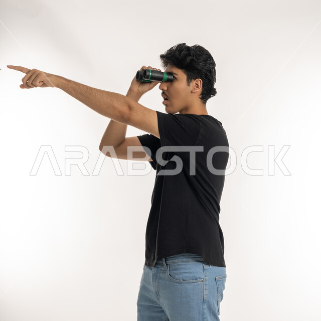 التخطيط المستقبلي للأهداف والطموحات، الإشارة بإصبع السبابة إلى مكان بعيد، حب المغامرة والاستكشاف، بورتريه لشاب عربي خليجي إماراتي يرتدي الزي الكاجوال ينظر من خلال المنظور ثنائي العينين (التلسكوب) بإيماءات الفرح والسرور، خلفية بيضاء