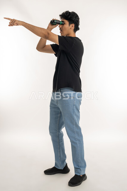 الإشارة بإصبع السبابة إلى مكان بعيد، حب المغامرة والاستكشاف، بورتريه لشاب عربي خليجي إماراتي يرتدي الزي الكاجوال ينظر من خلال المنظور ثنائي العينين (التلسكوب) بإيماءات الفرح والسرور، التخطيط المستقبلي للأهداف والطموحات، الطول الكامل للجسم، خلفية بيضاء