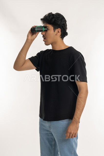 التخطيط المستقبلي للأهداف والطموحات، بورتريه لشاب عربي خليجي إماراتي يرتدي الزي الكاجوال ينظر من خلال منظار الرؤية, رؤية الاشياء البعيدة عبر المنظار، حب المغامرة والاستكشاف، ايماءات الفرح والاندماج, خلفية بيضاء