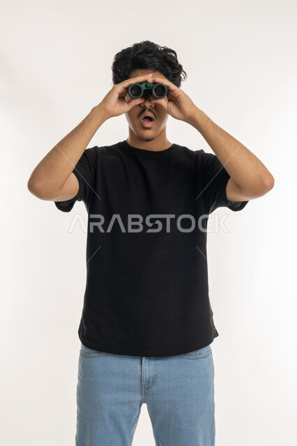 البحث عن شيء ما وتوسيع مجال الرؤية باستخدام المنظار، مفهوم الاستكشاف والمغامرة، رؤى مستقبلية عند الشباب، بورتريه لشاب عربي خليجي إماراتي يرتدي الكندورة يقوم بالنظر من خلال المنظار ثنائي العينين، إيماءات الاستغراب والمفاجأة، خلفية بيضاء