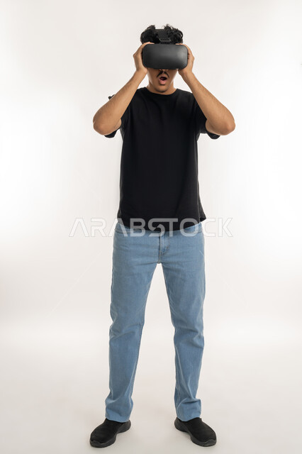 استخدام الأجهزة التقنية الحديثة والتكنولوجيا في رؤية المشاهد الخيالية، قضاء أوقات الفراغ بشكل ممتع، التفاعل والاندماج مع الواقع الافتراضي، بورتريه لشاب عربي خليجي إماراتي يرتدي الزي الكاجوال يضع نظارات الفي آر، الطول الكامل للجسم، خلفية بيضاء