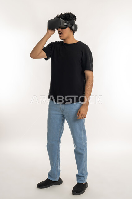 التفاعل والاندماج مع الواقع الافتراضي، بورتريه لشاب عربي خليجي إماراتي يرتدي الزي الكاجوال يضع نظارات الفي آر، استخدام الأجهزة التقنية الحديثة والتكنولوجيا في رؤية المشاهد الخيالية، قضاء أوقات الفراغ بشكل ممتع، الطول الكامل للجسم، خلفية بيضاء
