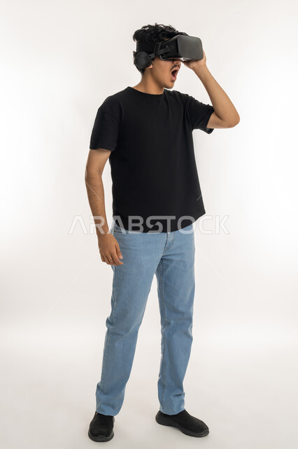 إيماءات التفاعل والاندماج مع الواقع الافتراضي، بورتريه لشاب عربي خليجي إماراتي يرتدي الزي الكاجوال يضع نظارات الفي آر، استخدام الأجهزة التقنية الحديثة والتكنولوجيا في رؤية المشاهد الخيالية، قضاء أوقات الفراغ بشكل ممتع، الطول الكامل للجسم، خلفية بيضاء