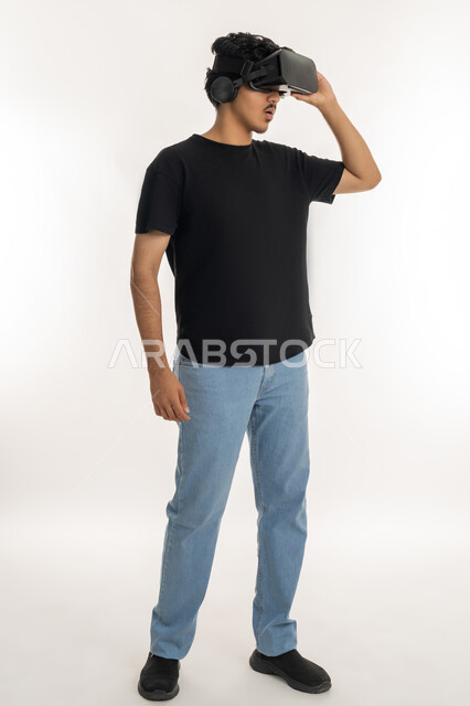 محاكاة العالم الخيالي والتفاعل مع الفيلم، تعابير وايماءات تدل على الاندماج والمتعة, بورتريه لشاب عربي خليجي إماراتي يرتدي الزي الكاجوال يقوم بمشاهدة أفلام عبر نظارات الواقع الافتراضي ثلاثية الأبعاد, الطول الكامل للجسم، خلفيه بيضاء