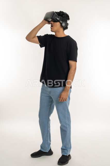 تعابير وايماءات تدل على الاندماج والمتعة, بورتريه لشاب عربي خليجي إماراتي يرتدي الزي الكاجوال يقوم بمشاهدة أفلام عبر نظارات الواقع الافتراضي ثلاثية الأبعاد, محاكاة العالم الخيالي والتفاعل مع الفيلم، الطول الكامل للجسم، خلفيه بيضاء