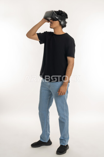قضاء أوقات الفراغ بشكل ممتع، إيماءات التفاعل والاندماج مع الواقع الافتراضي، بورتريه لشاب عربي خليجي إماراتي يرتدي الزي الكاجوال يضع نظارات الفي آر، استخدام الأجهزة التقنية الحديثة والتكنولوجيا في رؤية المشاهد الخيالية، الطول الكامل للجسم، خلفية بيضاء