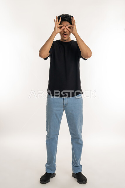 مفهوم التخطيط و الرؤية المستقبلية، بورتريه لشاب عربي خليجي اماراتي يرتدي الزي الكاجوال يضع يديه على عينيه على شكل نظارات بإيماءات التركيز، البحث والاستكشاف على شيء محدد، محاولة تقريب الأشياء البعيدة, الطول الكامل للجسم، خلفية بيضاء