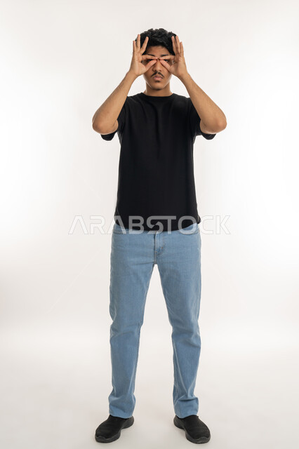 Trying to bring distant things closer, concept of planning and future vision, Portrait of a young Arab Gulf Emirati man wearing casual clothes putting his hands on his eyes in the form of glasses with gestures of concentration, searching and exploring a specific thing, full body length, white background