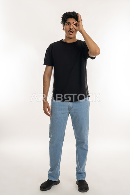 تعابير المشاهدة والتوضيح، بورتريه لشاب عربي خليجي إماراتي ينظر يضع يده على عينه بشكل عدسة بإيماءات الدهشة والذهول، الشعور بالفرح والسعادة، التدقيق والنظر إلى شيء محدد، مفهوم الرؤية المستقبلية، الطول الكامل للجسم، خلفية بيضاء