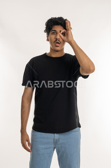 مفهوم الرؤية المستقبلية، تعابير المشاهدة والتوضيح، بورتريه لشاب عربي خليجي إماراتي ينظر يضع يده على عينه بشكل عدسة بإيماءات الدهشة والذهول، الشعور بالفرح والسعادة، التدقيق والنظر إلى شيء محدد، خلفية بيضاء