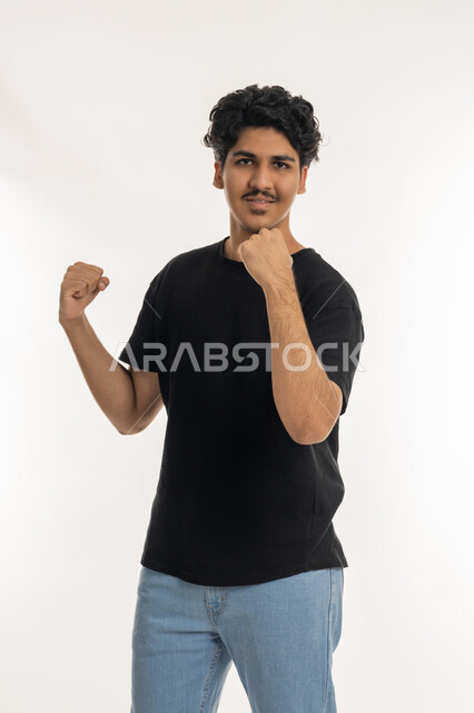 Gestures of happiness, victory and triumph, feeling of strength and success, expressions of interaction and integration, portrait of a young Arab Gulf Emirati man wearing casual clothes raising his hands with his palms together, clubs and cheering for the favorite team, white background