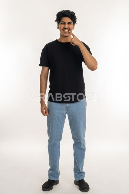 استخدام قشور فينير من الخزف لصنع ابتسامة بيضاء ناصعه، أهمية متابعة طبيب الاسنان، العناية والتجميل والتبيض، بورتريه لشاب عربي خليجي إماراتي يرتدي الزي الكاجوال ينظر الى الكاميرا يشير بيديه إلى أسنانه بإيماءات السرور، الطول الكامل للجسم، خلفية بيضاء