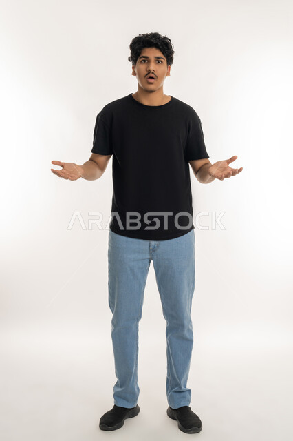 Surprise and inquiry about something, questioning about a specific topic, portrait of a young Arab Gulf Emirati man wearing casual clothes looking at the camera and raising his hands up with gestures of uncertainty, feeling confused, vague and unclear, full body length, white background