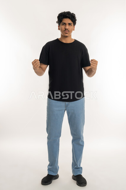 Trying to control oneself and oneself, strength and ability to do something, expressions of anger, annoyance and defiance, portrait of a young Arab Gulf Emirati man wearing casual clothes raising his fists in excitement, full body length, white background