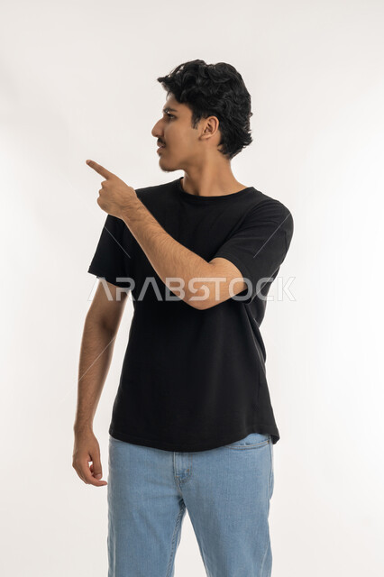 الإشارة بأصبع السبابة الى هدف محدد، ، بورتريه لشاب عربي خليجي إماراتي مبتسم يرتدي الزي الكاجوال ينظر الى الكاميرا و يرفع يده للأعلى لتحديد موقع معين، مفهوم النقر والضغط والتفاعل مع العناصر الهولوجرامية، خلفية بيضاء