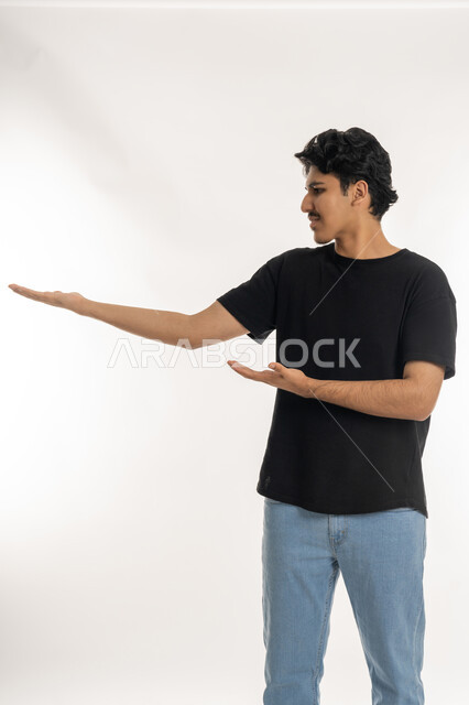 النظر الى شيء ما بتعابير الفرح والسعادة، الترحيب والاستقبال بحب وبشاشة، بورتريه لشاب عربي خليجي إماراتي مبتسم يرتدي الزي الكاجوال يفتح كفيه بإيماءات السرور برؤية شخص ما، خلفية بيضاء