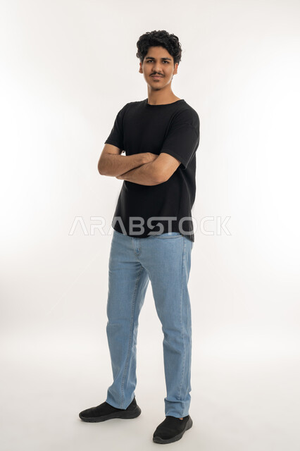 The concept of elegance and chic, interest in external appearance, side portrait of a young Arab Gulf Emirati man wearing casual clothes standing with crossed hands, looking at the camera with gestures of happiness and pleasure, expressions of masculinity and self-confidence, full body length, white background