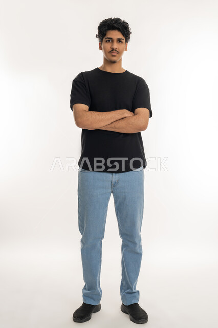 ايماءات الثقة بالنفس، مفهوم الاهتمام بالمظهر الخارجي و الرجولة والأناقه، بورتريه لشاب عربي خليجي إماراتي يرتدي الزي الكاجوال يقف باستقامة بايدي متقاطعه, النظر إلى الكاميرا بتعابيرالجدية والحزم، صورة الجسم بالطول الكامل، خلفية بيضاء