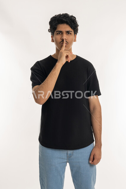 الهدوء والسكون وعدم الإزعاج، التوقف عن الكلام وخفض الصوت، بورتريه لشاب عربي خليجي إماراتي يرتدي الزي الكاجوال يضع أصبع السبابة على فمه بإيماءات طلب السكوت والصمت، النظر إلى الكاميرا بتعابير الغضب، الطول الكامل للجسم, خلفية بيضاء