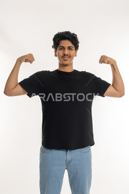 بناء جسم رياضي، المحافظة على اللياقه البدنيه، بورتريه لشاب عربي  خليجي إماراتي يرتدي الزي الكاجوال يرفع يديه نحو الأعلى وينظر الى  الكاميرا بايماءات القوة والافتخار, حركة العضلات ذات الرأسين, خلفية بيضاء