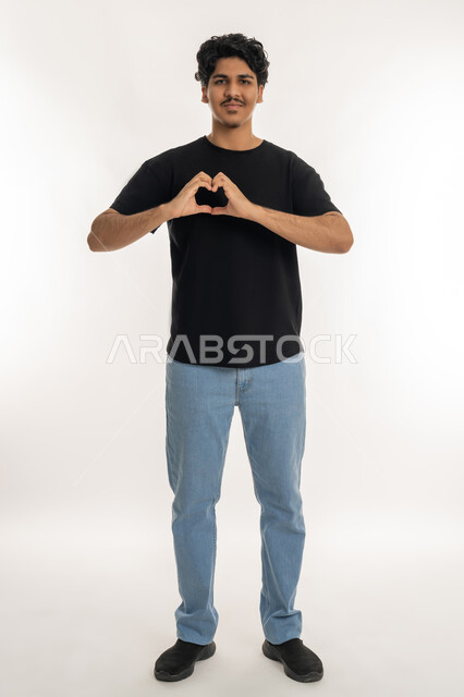 مفهوم المحبة والمودة، ايماءات يدين على شكل قلب،, بورتريه لشاب عربي خليجي إماراتي مبتسم يرتدي الزي الكاجوال ينظر إلى الكاميرا بتعابير السعادة والفرح, اظهار مشاعر الحب والشغف, الطول الكامل للجسم، خلفية بيضاء