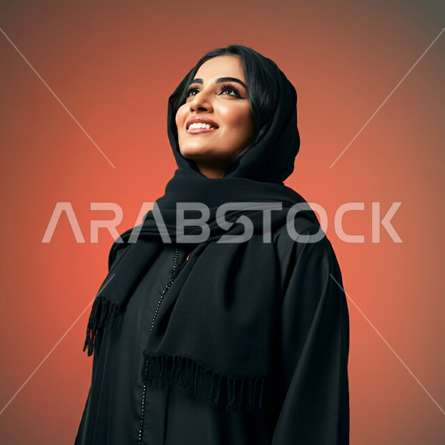 مفهوم الأنوثة والنعومة، بورتريه مقرب من الجانب لامرأة عربية خليجية سعودية محجبة مبتسمة ترتدي العباءة السوداء تنظر إلى الأعلى  بإيماءات الثقة بالنفس والفرح، الاهتمام بالأناقة والمظهر الخارجي، تعابير السرور والسعادة، خلفية ملونة