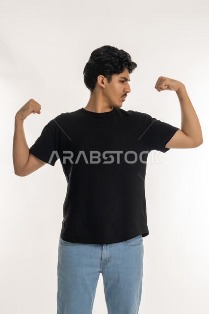 حركة العضلات ذات الرأسين, بناء جسم رياضي، المحافظة على اللياقه البدنيه، بورتريه لشاب عربي  خليجي إماراتي يرتدي الزي الكاجوال يرفع يديه نحو الأعلى وينظر الى عضلاته, إيماءات وجه ويدين تدل على القوه و الصحه الجسديه, خلفية بيضاء