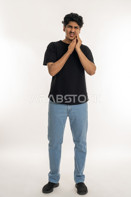 الإحساس بوجع الأسنان والتألم من النخور، الحاجة للذهاب الى العيادة السنية لوضع الخطة العلاجية، الشعور بالتعب والمرض, بورتريه لشاب عربي خليجي إماراتي يرتدي الزي الكاجوال يضع يديه على خده بتعابير الألم, تسوس الأضراس وإلتهاب اللثه, الجسم بالطول الكامل، خلفية بيضاء