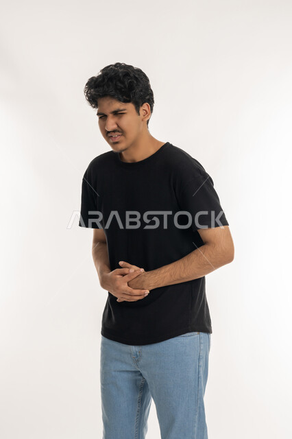 Facial and hand movements indicating disturbance and discomfort, the need for rest and appropriate treatment, portrait of a young Arab Gulf Emirati man wearing casual clothes, putting his hands on his stomach with expressions of pain, feeling sick and stomach pain, white background