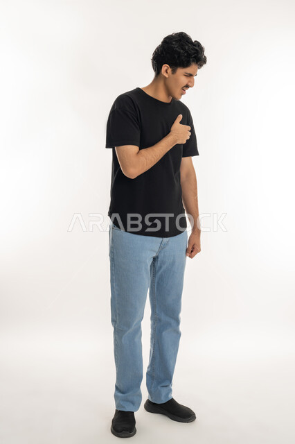 الشعور بحالة سيئة من الوخز والألم, اضطرابات وأمراض قلبية, التعب والإرهاق والضغوطات النفسية, ضرورة الذهاب للطبيب لتلقي العلاج المناسب، الاحساس بذبحه صدريه، بورتريه لشاب عربي خليجي إماراتي يرتدي الزي الكاجوال يضع يده على صدره، الطول الكامل للجسم، خلفية بيضاء