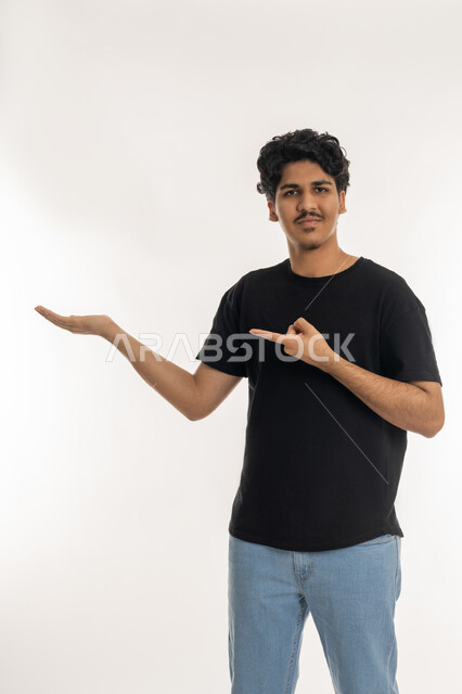 النظر إلى الكاميرا بإيماءات البشاشة, إمساك شيء معين في كف اليد، بورتريه لشاب عربي خليجي إماراتي يرتدي الزي الكاجوال يحمل عنصر ما ويشير إليه بإصبع السبابة، خلفية بيضاء