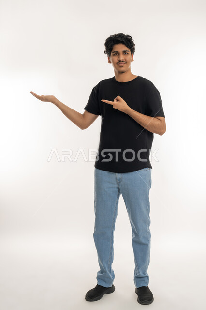 الوقوف والنظر إلى الكاميرا بإيماءات البشاشة, إمساك شيء معين في كف اليد، بورتريه لشاب عربي خليجي إماراتي يرتدي الزي الكاجوال يحمل عنصر ما ويشير إليه بإصبع السبابة، الطول الكامل للجسم, خلفية بيضاء