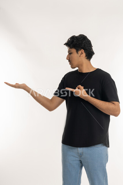 إمساك شيء معين في كف اليد، الإشارة إلى عنصرما بإصبع السبابة، بورتريه لشاب عربي خليجي إماراتي يرتدي الزي الكاجوال يقف باستقامة، النظر إلى يده بايماءات الجدية والحزم، الخلفية بيضاء