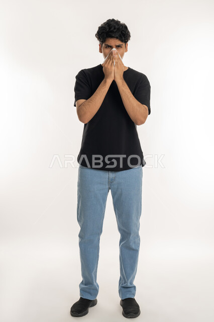 Need for treatment and medication, allergic rhinitis and sinusitis, feeling sick and cold, portrait of a young Arab Gulf Emirati teenager in casual wear putting a tissue on his face with sneezing gestures, full body, white background