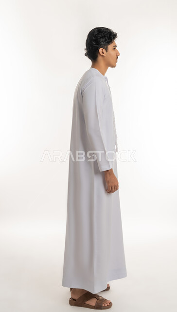 Concern for appearance, concept of elegance and masculinity, side portrait of a young Arab Gulf Emirati teenager wearing a kandura standing straight and upright, full body, white background