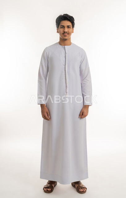 Concept of elegance and masculinity, expressions of self-confidence, interest in appearance, portrait of a young Arab Gulf Emirati teenager wearing a kandura standing straight and looking at the camera with gestures of self-pride, full-length body, white background