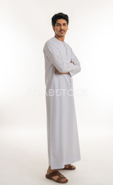 Concern about appearance, concept of elegance and masculinity, expressions of self-confidence, portrait of a young Arab Gulf Emirati teenager wearing a kandura standing straight with crossed hands and looking at the camera with gestures of self-esteem, full body, white background