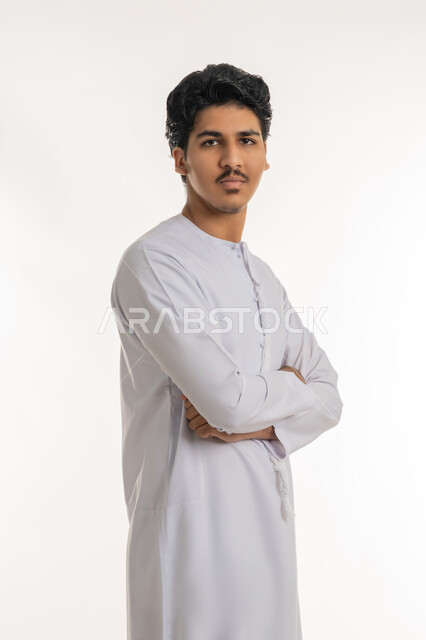الاهتمام بالمظهر الخارجي, مفهوم الأناقة و الرجولة, تعابير الثقة بالنفس, بورتريه لشاب مراهق عربي خليجي إماراتي يرتدي الكندورة يقف باستقامة وأيدي متقاطعة وينظر إلى الكاميرا بإيماءات الاعتزاز بالذات, خلفية بيضاء