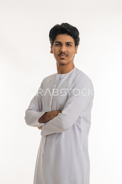 Concern about appearance, concept of elegance and masculinity, expressions of self-confidence, portrait of a young Arab Gulf Emirati teenager wearing a kandura standing straight with crossed hands and looking at the camera with gestures of self-esteem, white background